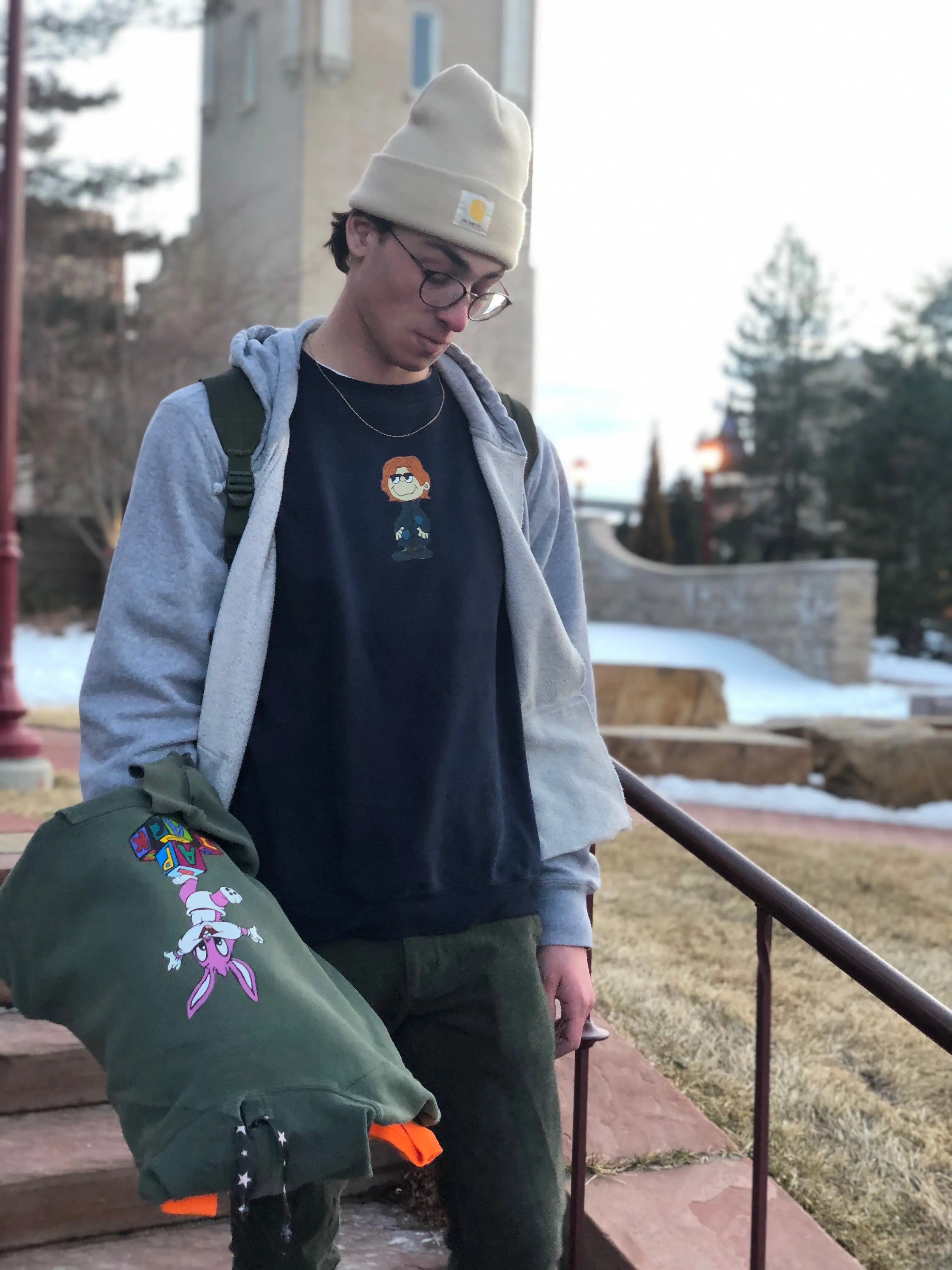 Person wearing a gray hoodie, black t-shirt with a graphic, and green jacket with colorful designs outdoors.